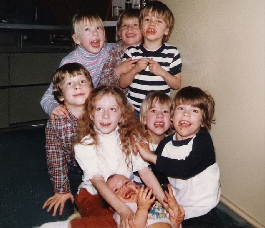 A group of seven smiling children sit closely together on the floor, with one baby lying on their laps. The kids are laughing and looking at the camera against a plain wall background.