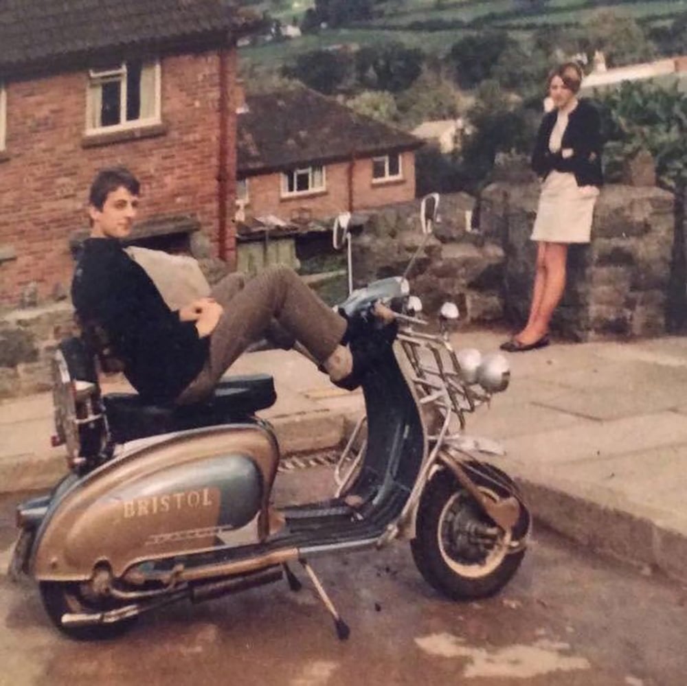 A young man leans back on a vintage BRISTOL scooter parked on a driveway, while a young woman in a light skirt and dark jacket stands nearby, a stone wall and houses visible in the background.