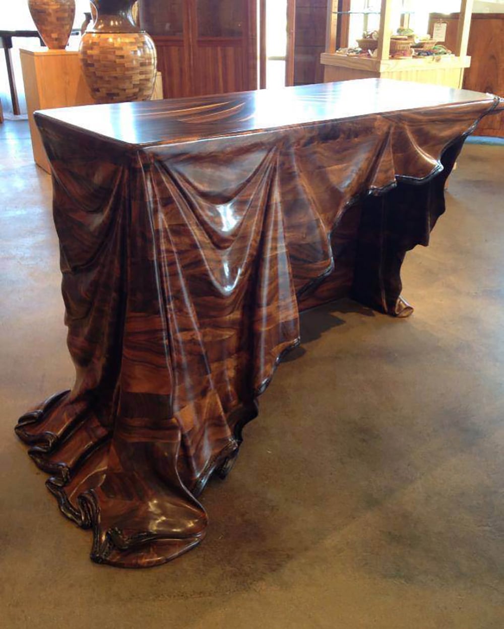 A wooden table designed to look like it is draped with a flowing, detailed cloth, with realistic folds and creases carved into the wood, standing in a warmly lit room with wooden furniture in the background.