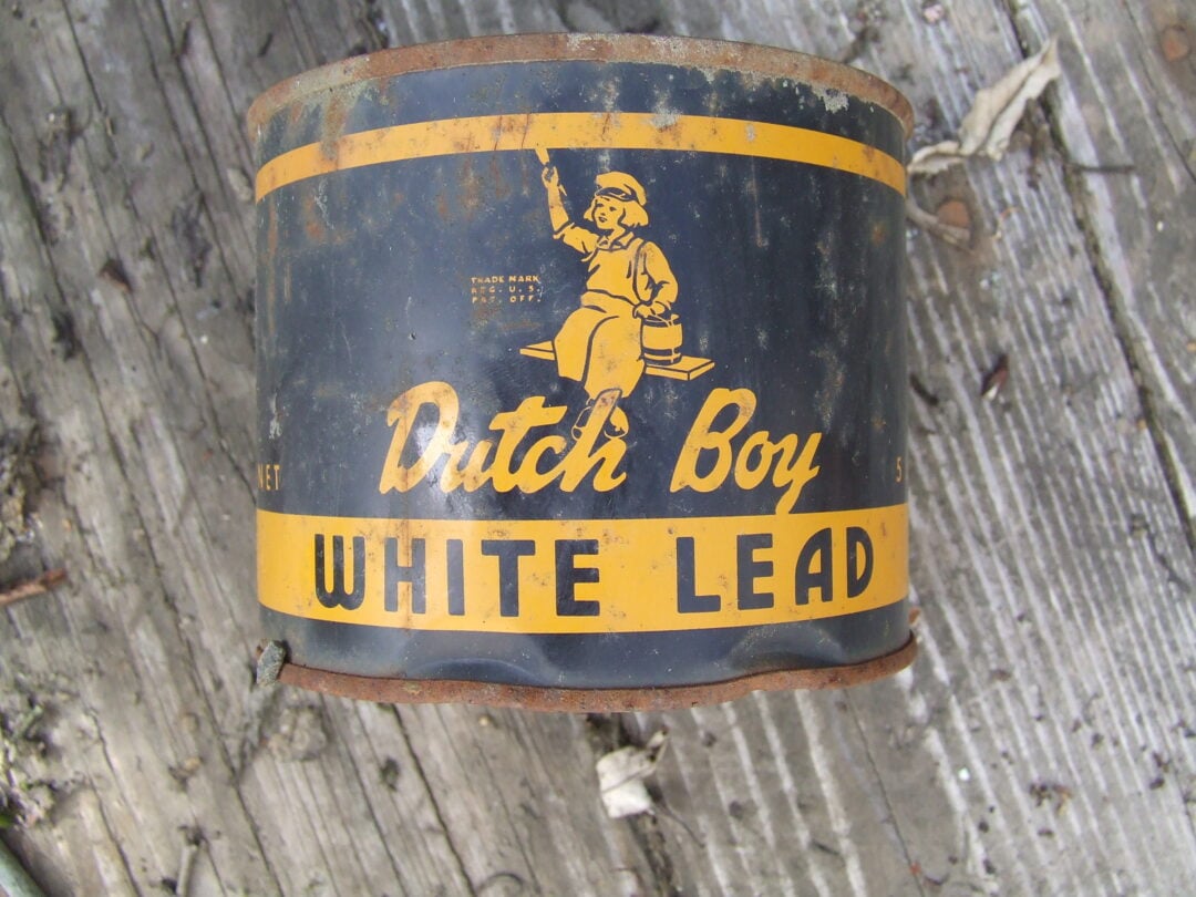 A rusty, vintage metal can labeled "Dutch Boy White Lead" with a yellow and navy design, featuring an illustration of a boy holding a paintbrush, sits on weathered wooden boards.