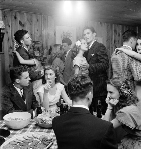 Black-and-white photo of teenagers at a party; some are dancing in pairs, while others sit at a table with snacks, chips, cookies, and soda bottles. The room has wood-paneled walls and a relaxed, vintage 1950s vibe.