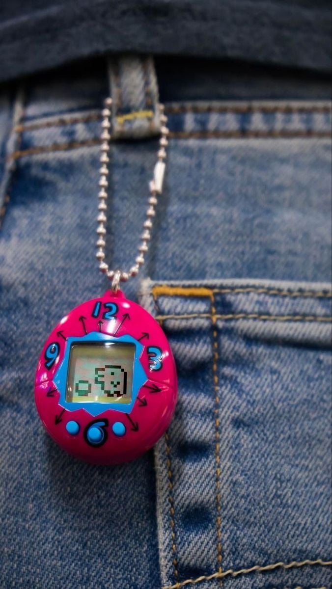 A pink digital pet keychain with a chain hangs over the front pocket of blue denim jeans. The device's screen displays a pixelated character.