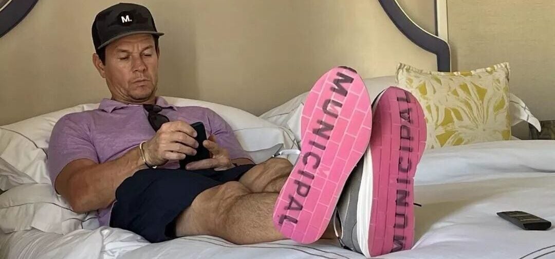 A man wearing a purple shirt, navy shorts, and a black cap relaxes on a bed, holding a phone. His shoes, with "MUNICIPAL" printed in pink on the soles, are propped up and visible. A pillow and a remote are nearby.
