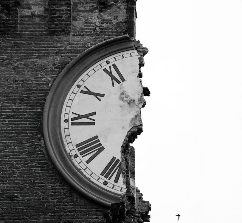 A large, old clock face is partially destroyed, with only half remaining on a brick wall. The missing portion reveals jagged edges, and the sky is visible in the background. A small bird is flying nearby.