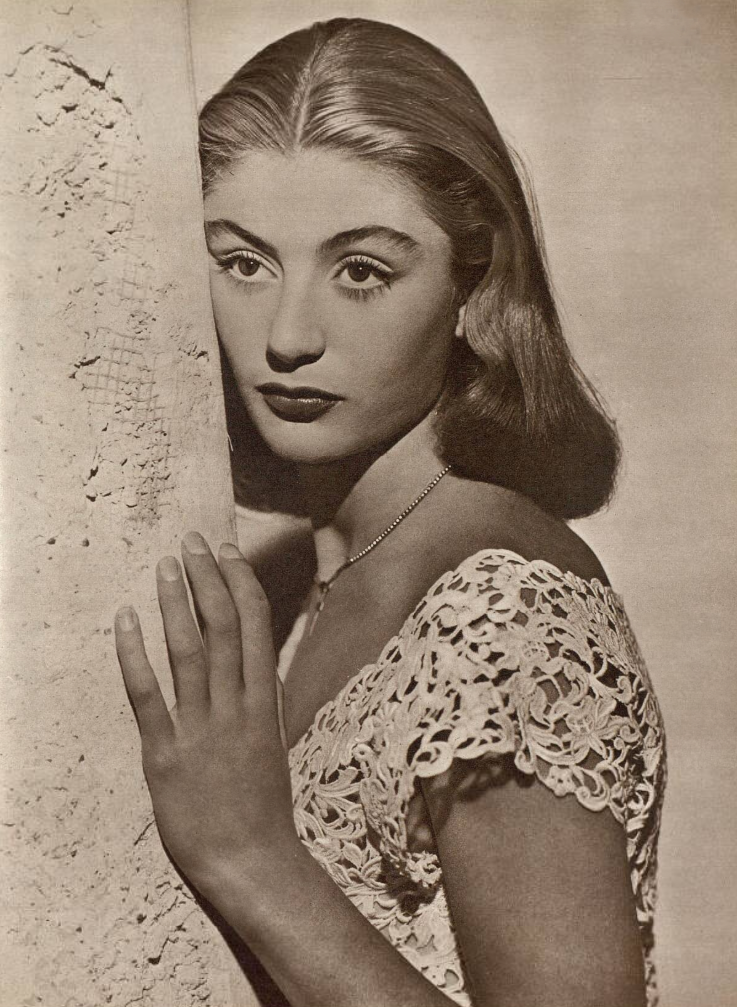 A young woman with wavy, shoulder-length hair and a lace dress leans against a textured wall, gazing thoughtfully into the distance in a black-and-white portrait.