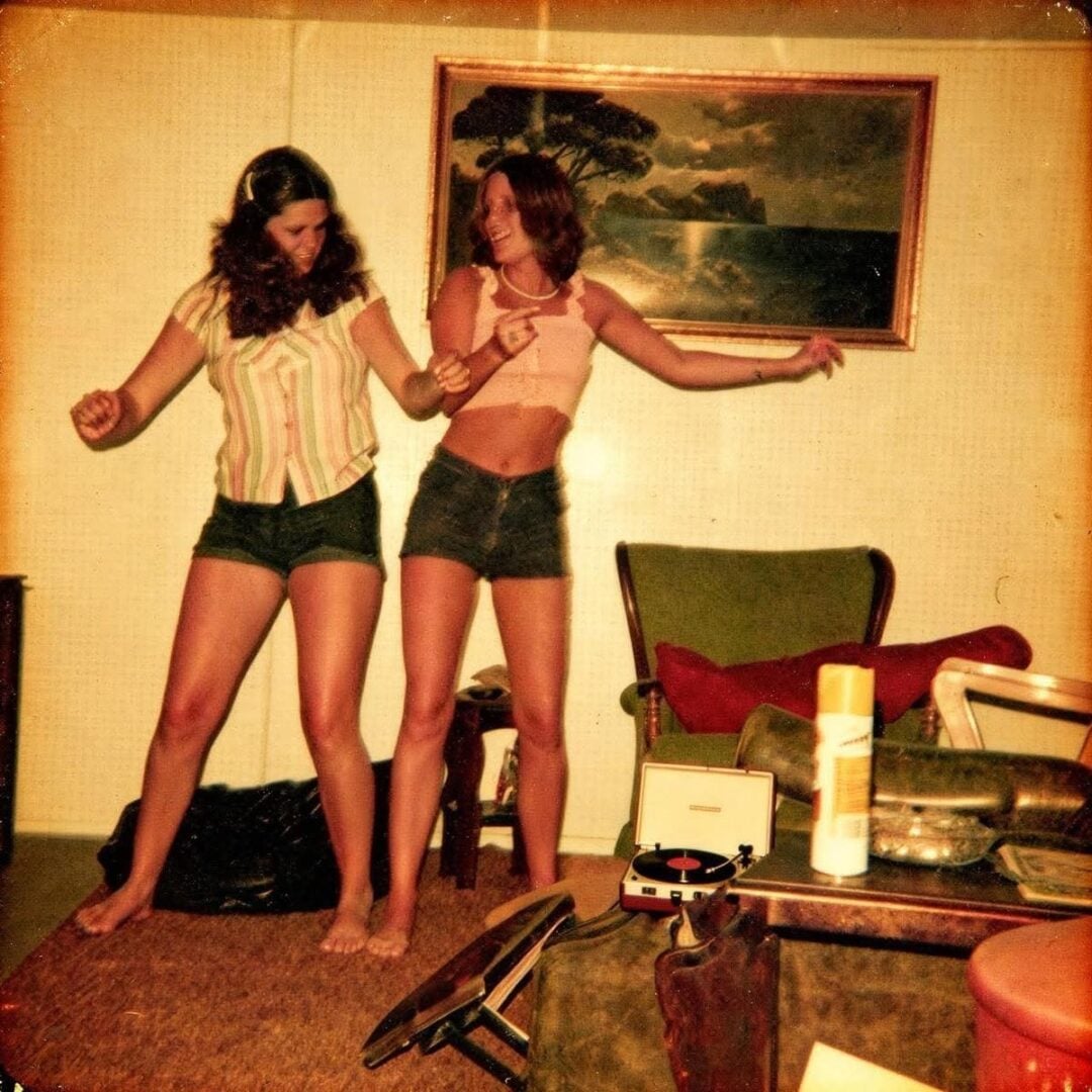 Two young women dance barefoot in a retro living room with vintage furniture. One wears shorts and a striped shirt, the other a tank top and shorts. A record player and a framed painting are visible in the background.
