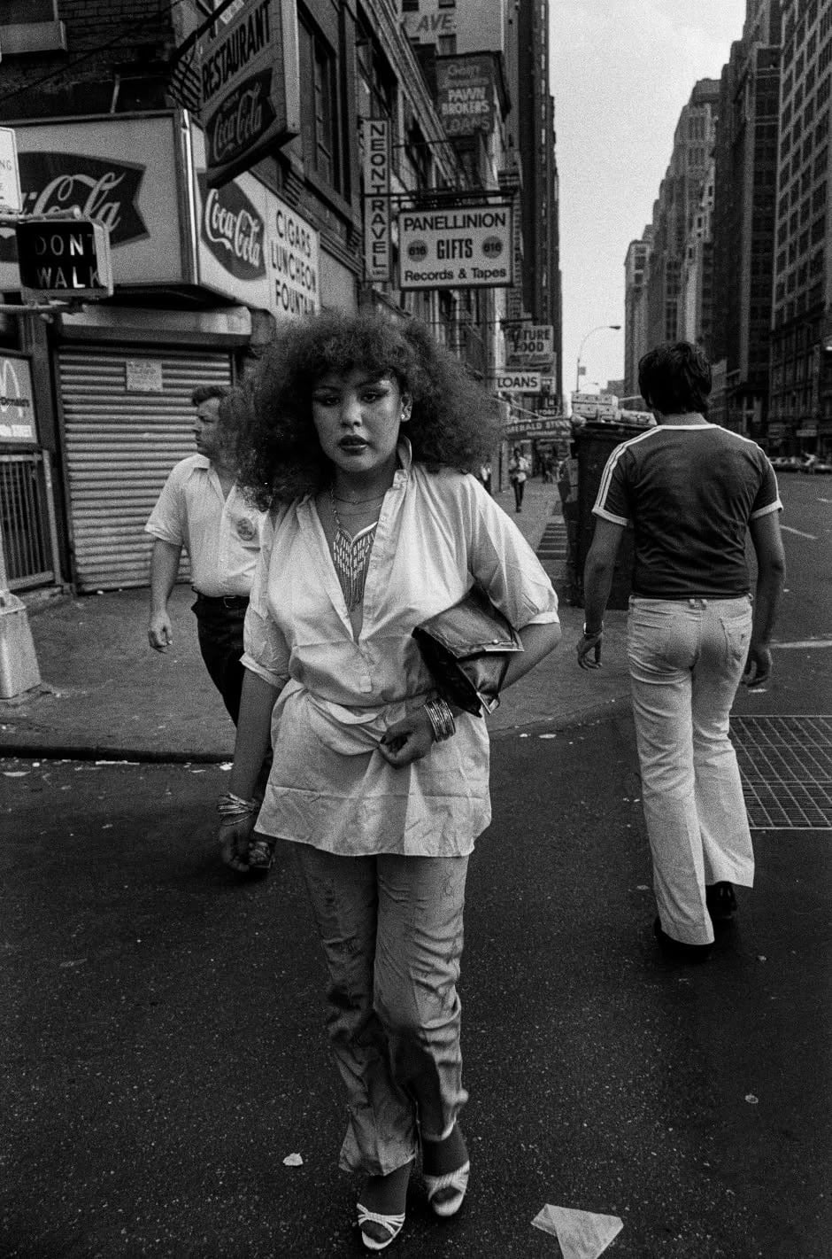 A woman with voluminous hair and a stylish outfit walks down a busy city street, holding a clutch. Several people walk nearby. Tall buildings, shop signs, and a Coca-Cola sign are visible in the background.