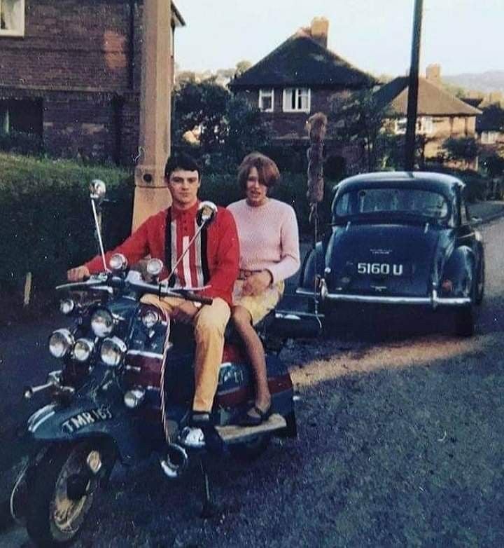 A young man and woman sit on a vintage scooter parked on a residential street, with a classic car and brick houses in the background. The man wears a red jacket; the woman is in a pink sweater and skirt.