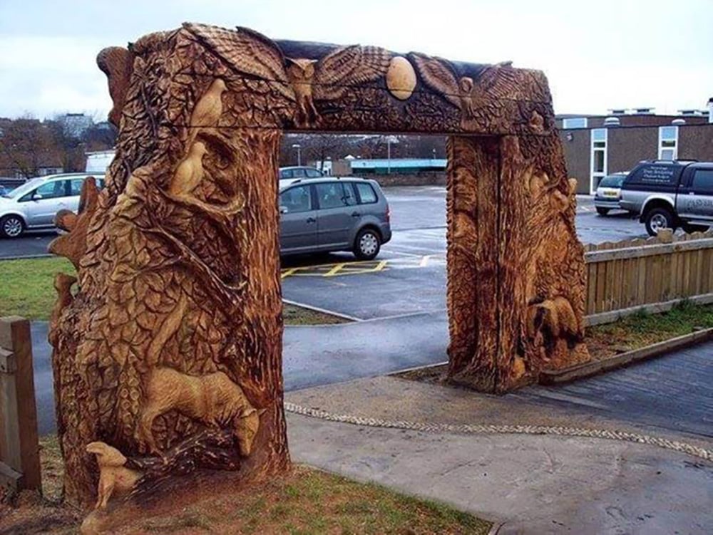 A large wooden archway intricately carved with forest animals, leaves, and trees stands beside a parking lot, with cars and buildings visible in the background.