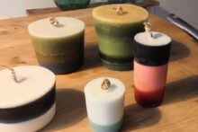 Five colorful pillar candles are arranged on a wooden table next to a saucepan filled with used tealight holders and candle scraps. Green glass vases and a mirror are in the background.