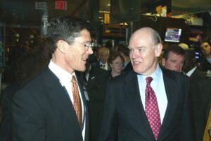 Two men in suits are talking and smiling while walking indoors; several other people, some in yellow jackets, are visible in the background. The setting appears to be a busy financial trading floor.