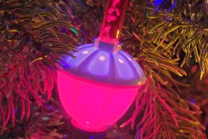 A close-up of a Christmas tree branch decorated with colorful lights, a glowing pink bubble light ornament, and a green bauble, surrounded by green pine needles.