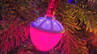 A close-up of a Christmas tree branch decorated with colorful lights, a glowing pink bubble light ornament, and a green bauble, surrounded by green pine needles.