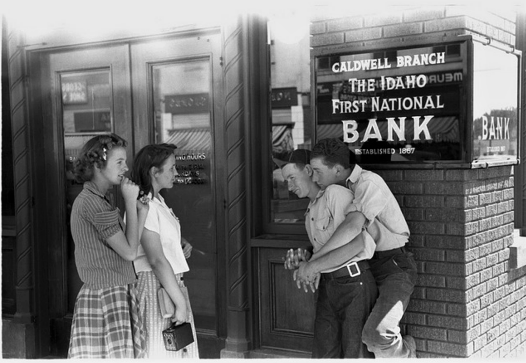 Four teenagers stand outside the Caldwell Branch of The Idaho First National Bank; two girls wait by the bank door while two boys playfully wrestle near a window displaying the bank’s sign.