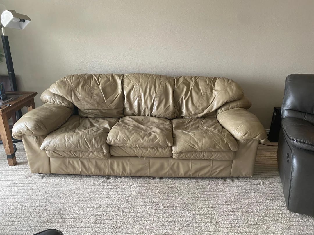 A worn beige leather sofa with three seat cushions sits on a light-colored carpet in a living room, next to a wooden table and a dark armchair.