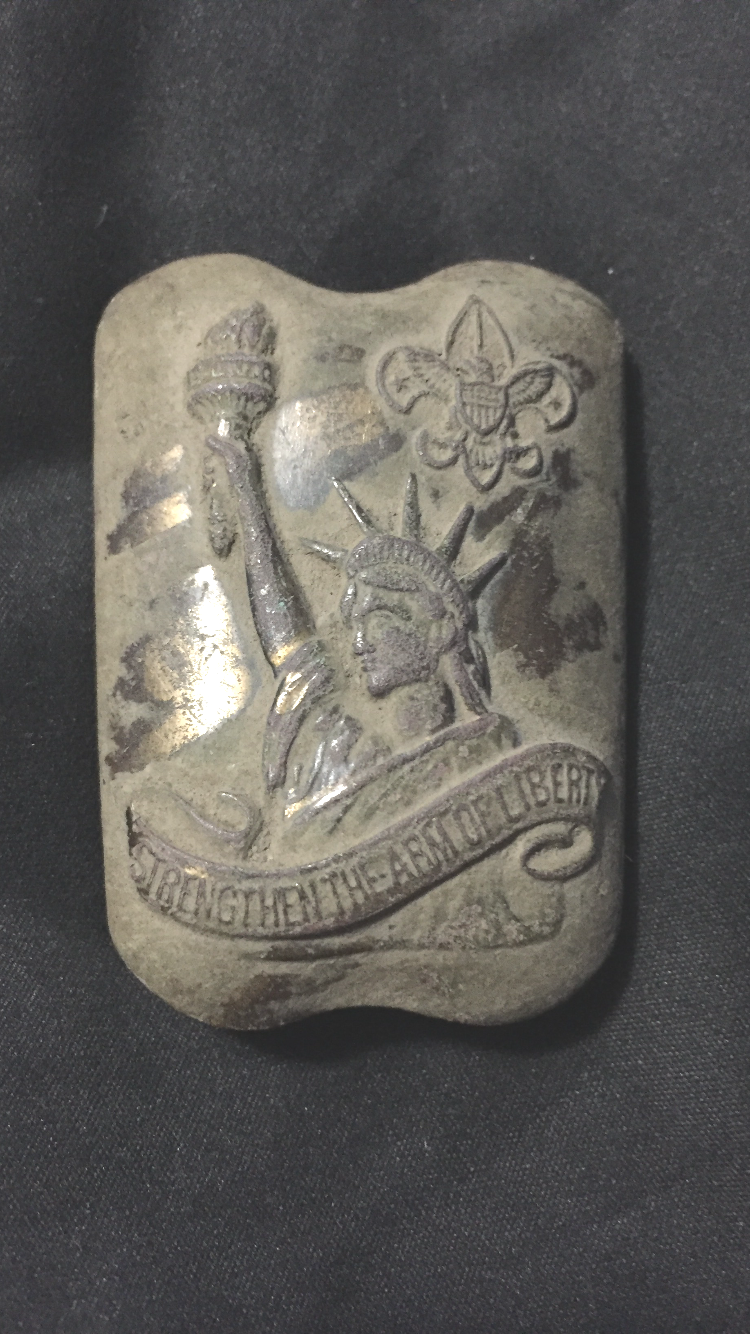 A metallic object with an engraving of the Statue of Liberty holding a torch, the Boy Scouts fleur-de-lis emblem above, and a ribbon reading “STRENGTHEN THE ARM OF LIBERTY,” on a dark fabric background.