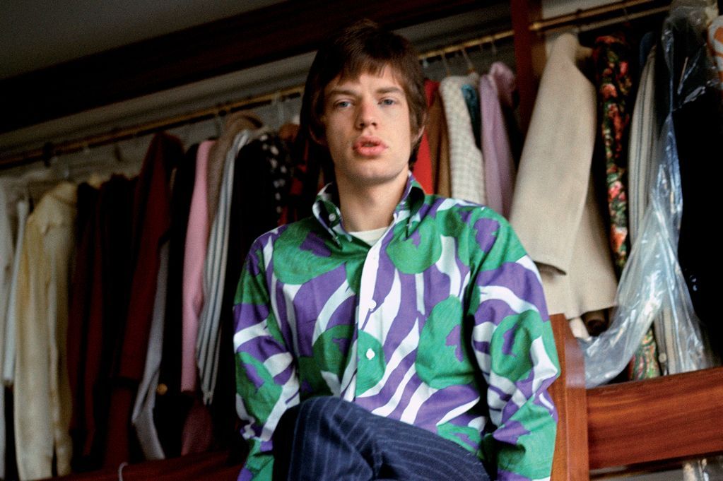 A young man with brown hair wearing a bold, colorful patterned shirt sits in front of an open wardrobe filled with assorted hanging clothes.