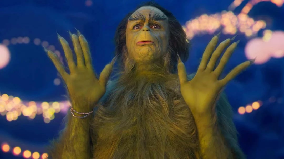 A fuzzy green character with yellow skin, long fingers, and expressive eyebrows holds up both hands, standing against a blurred background of colorful holiday lights.