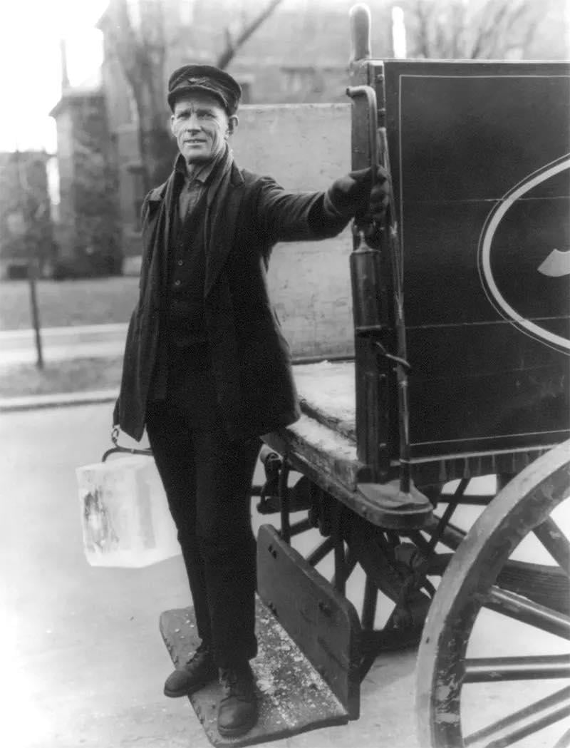 Black-and-white photo of a man in uniform holding a metal can and standing beside a horse-drawn wagon. The man wears a cap and coat and rests one arm on the wagon's side. Buildings and trees are in the background.