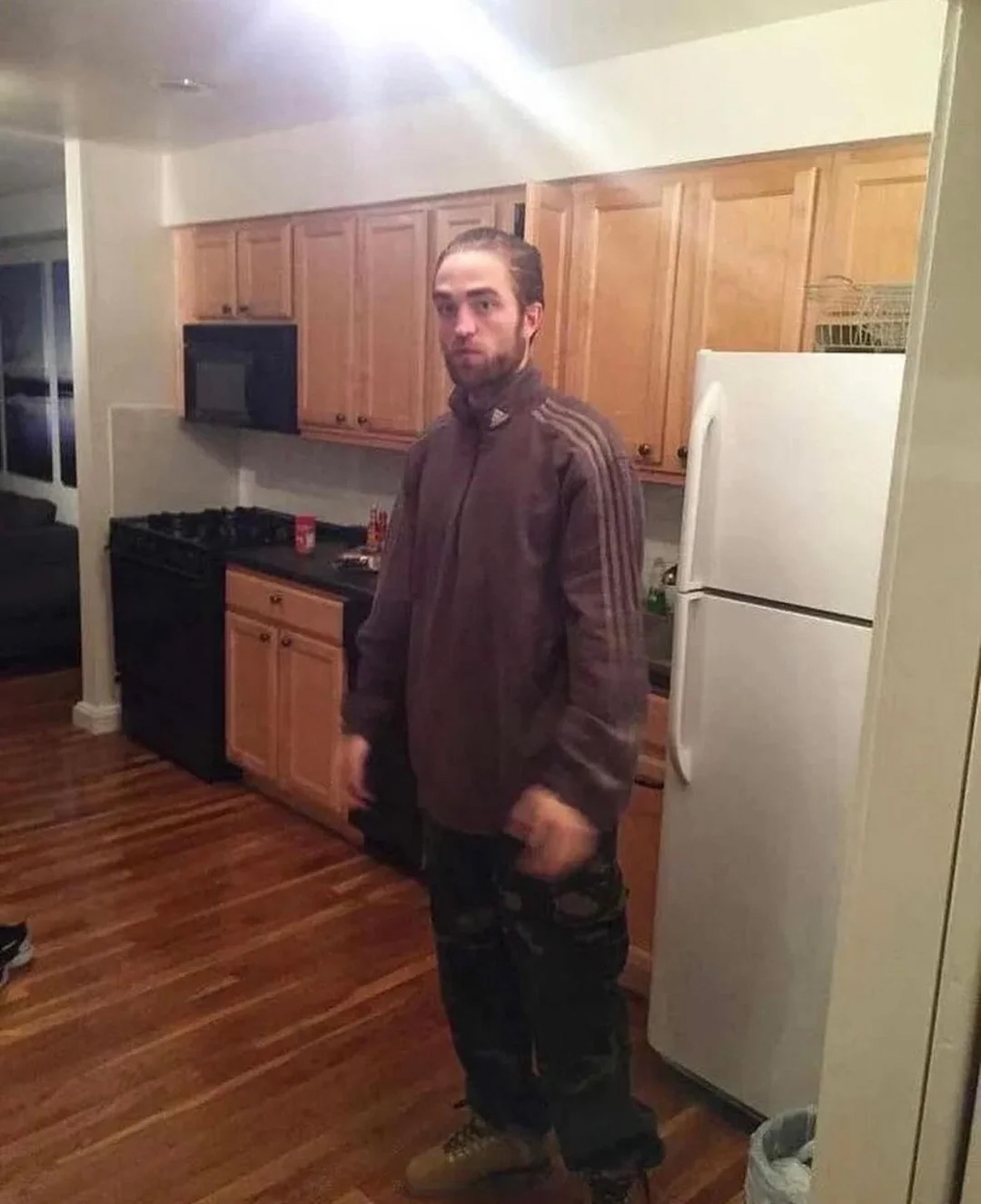 A man with a beard stands in a kitchen with wooden floors, light wood cabinets, a white refrigerator, and a black stove. He is wearing a brown jacket, dark pants, and tan boots.