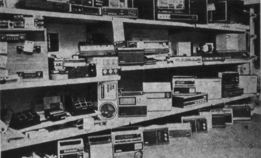 Black and white photo of several shelves filled with various vintage electronic devices, such as radios, cassette players, and audio equipment, arranged haphazardly.