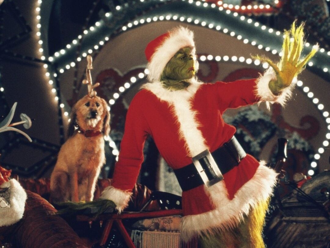 The Grinch, dressed in a red Santa suit, stands with one arm extended. His loyal dog, Max, with a fake antler tied to his head, sits nearby. The background is decorated with bright, festive holiday lights.