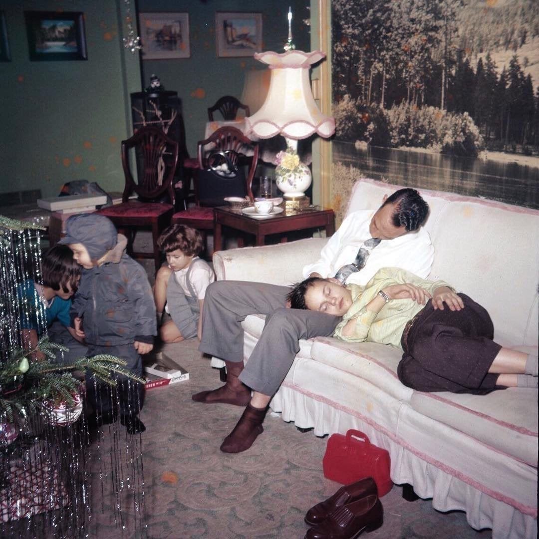 A man and a woman nap on a sofa in a living room, while children play nearby. The room has vintage decor, with a pink lamp, artwork, chairs, and a large mural of a forest on the wall.