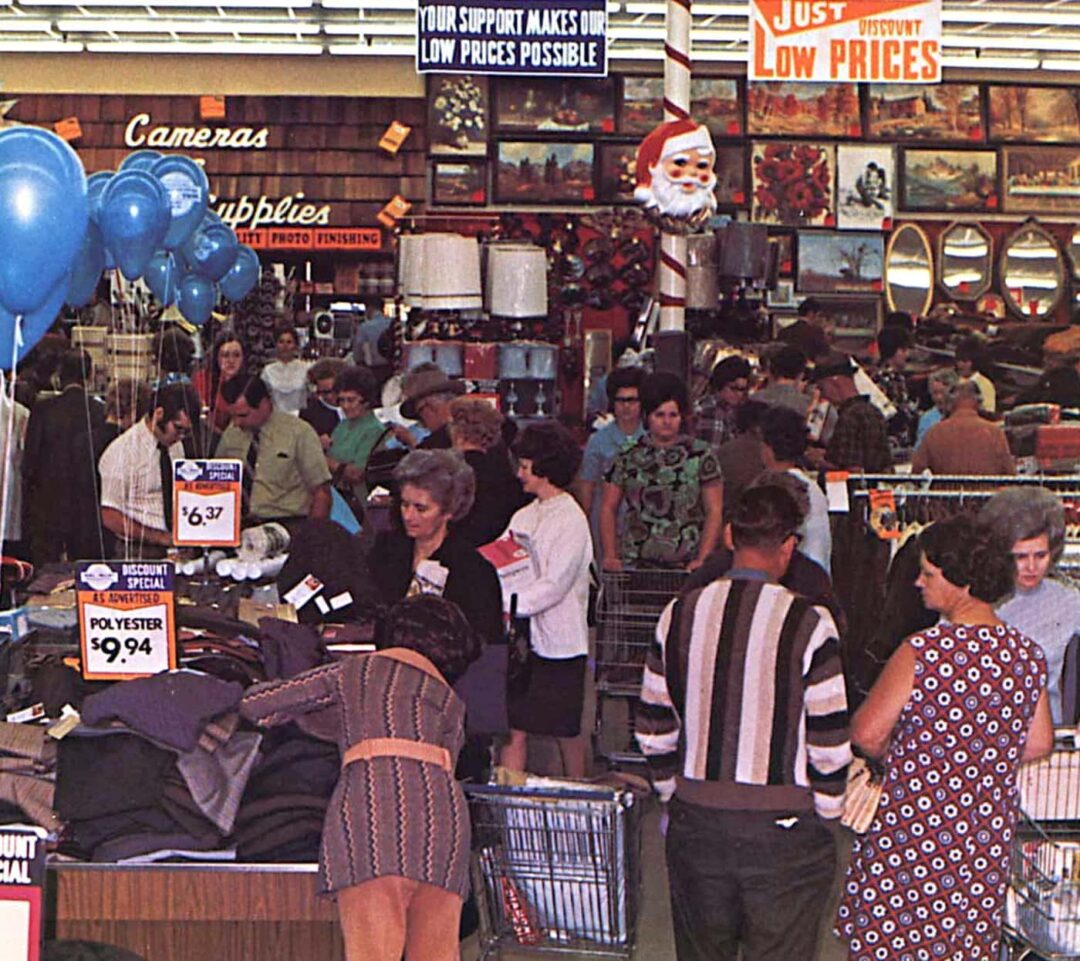 A busy 1970s department store with shoppers browsing clothing racks and signs advertising low prices; blue balloons, Santa decoration, and various items for sale are visible among the crowd.