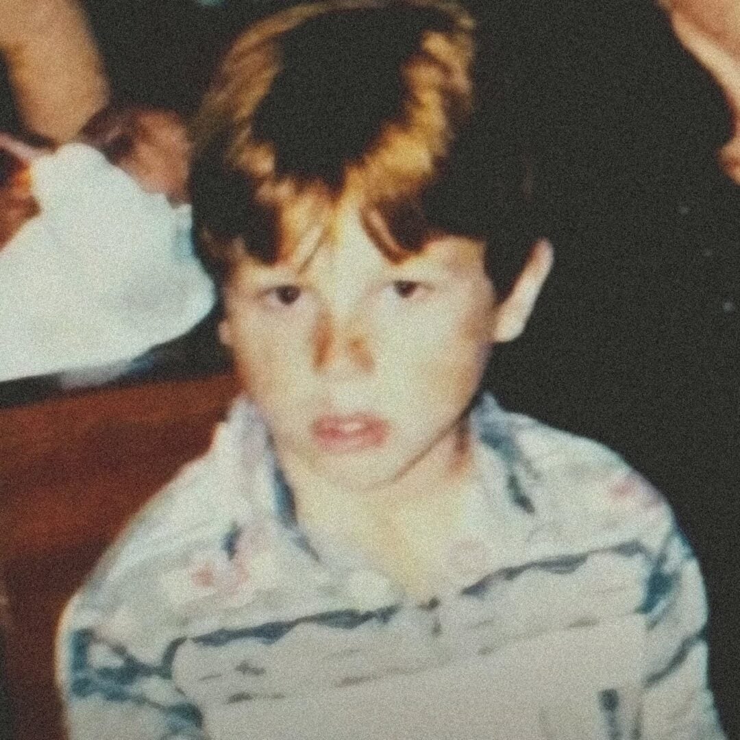 A young boy with light skin and reddish-brown hair is looking slightly past the camera. He is wearing a floral-patterned shirt, and the image has a blurry, vintage quality.