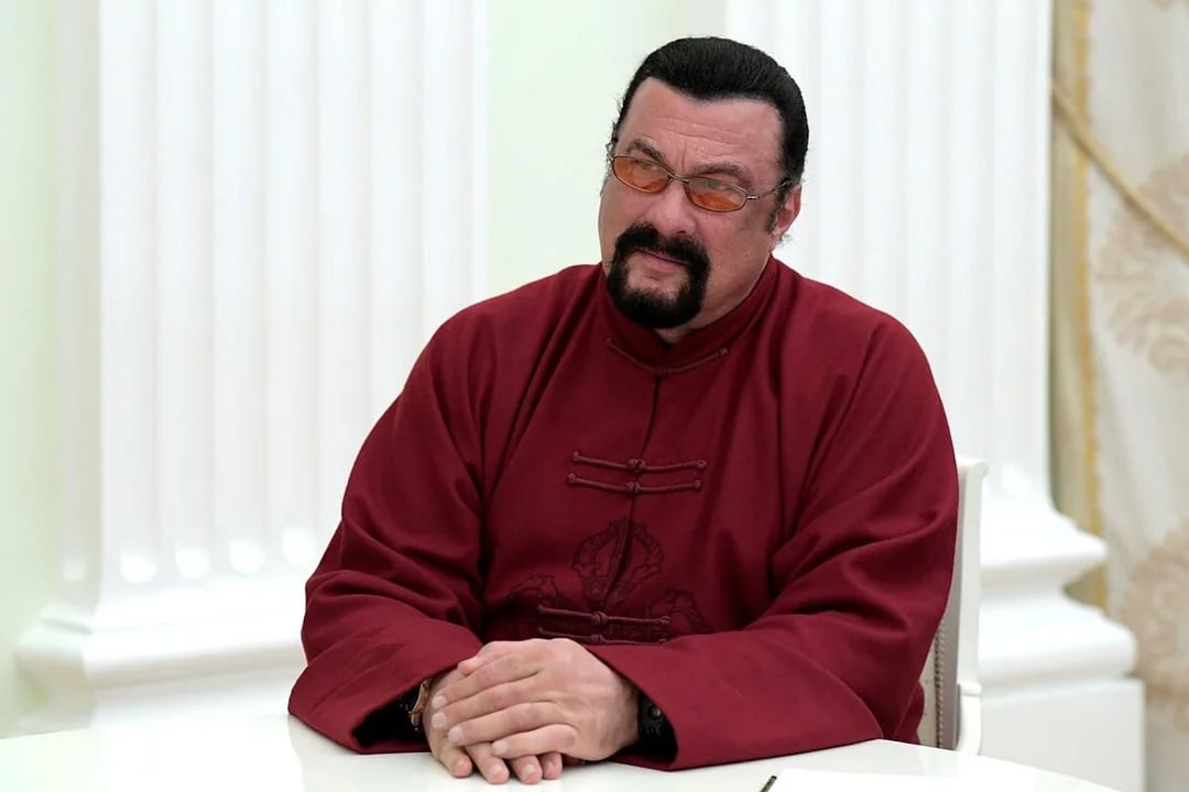 A man with dark hair, a beard, and mustache, wearing tinted glasses and a maroon traditional-style robe, sits at a white table with his hands folded, in a room with white columns and light-colored walls.
