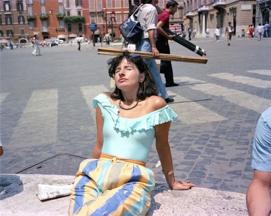 A woman in a mint green off-shoulder top and striped pants sits on a stone bench in a sunny city square, eyes closed. Behind her, a man appears to be balancing wooden planks on her head due to perspective.