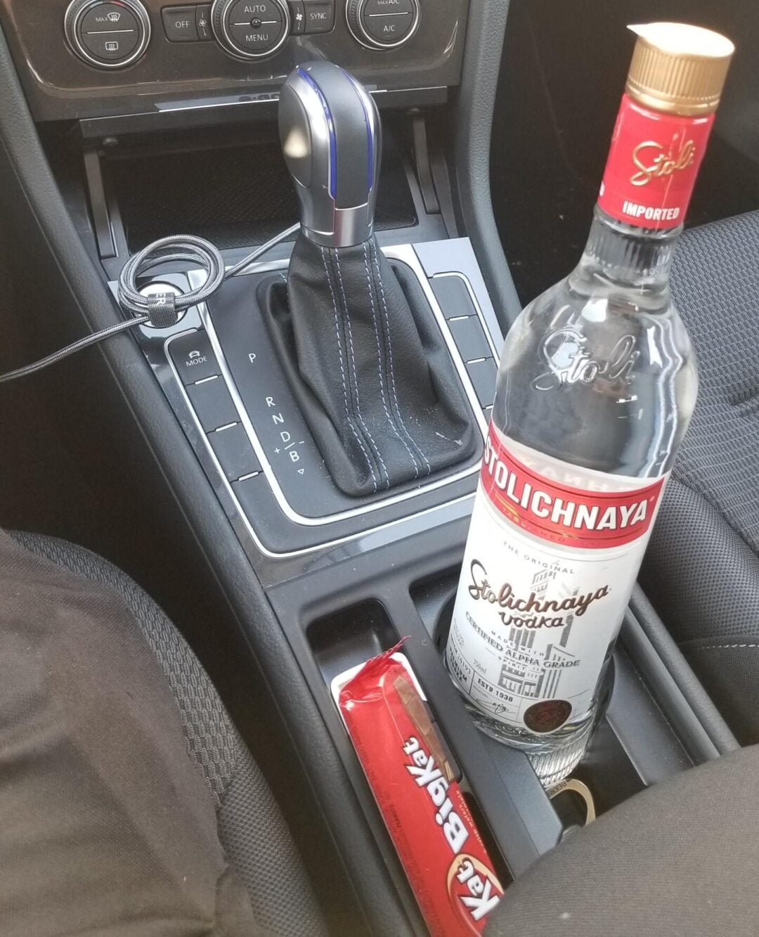 A car's center console with an automatic gear shift, a bottle of Stolichnaya vodka, an unopened chocolate bar, and a pair of wired earphones resting nearby.