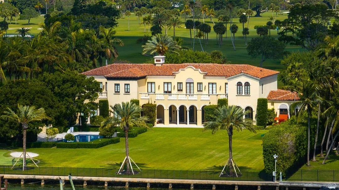 A large Mediterranean-style mansion with a red-tiled roof, arched windows, and balconies, surrounded by lush palm trees, manicured lawns, and a swimming pool near a waterfront.