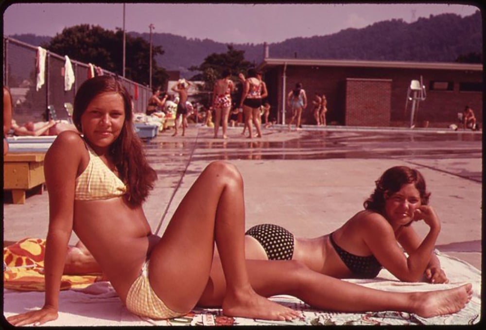 Two teenage girls in bikinis relax on towels by an outdoor public pool on a sunny day, with other swimmers and trees visible in the background. One girl sits upright while the other lies on her stomach, smiling at the camera.