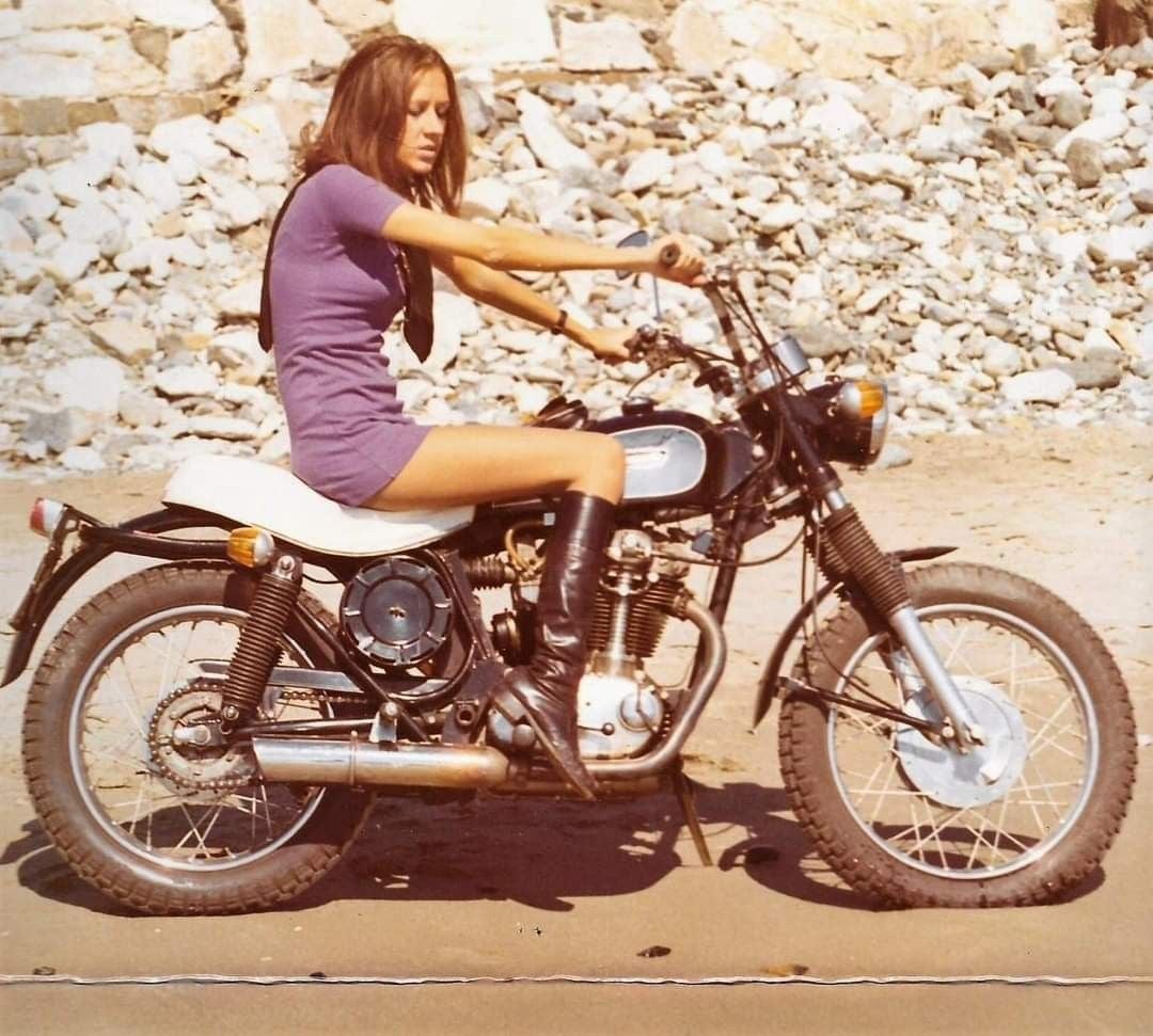 A woman in a purple dress and black boots sits on a vintage motorcycle parked on a sandy surface, with rocks and stones in the background.