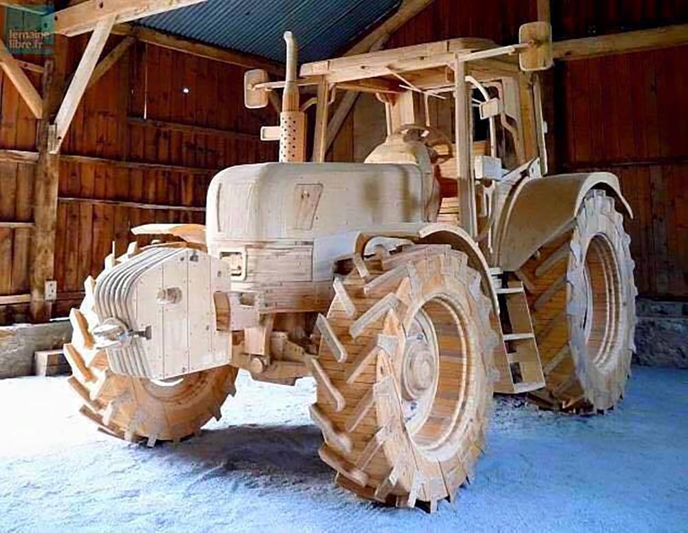 A life-sized tractor sculpture made entirely of wood stands inside a wooden barn, showcasing detailed craftsmanship, including textured tires, engine parts, and cabin.