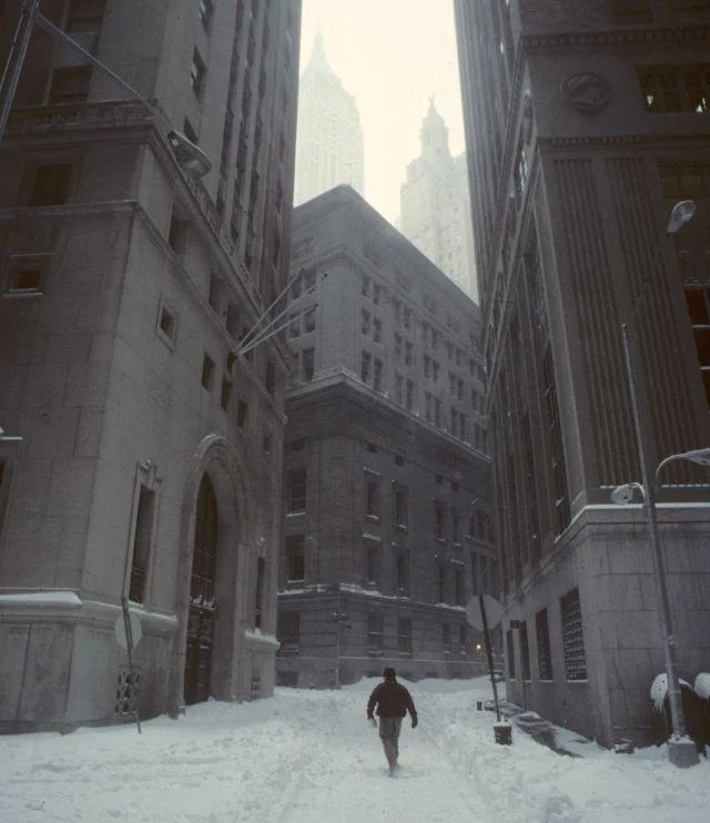 A person walks alone down a snow-covered street surrounded by tall, old buildings in a city on a foggy winter day.