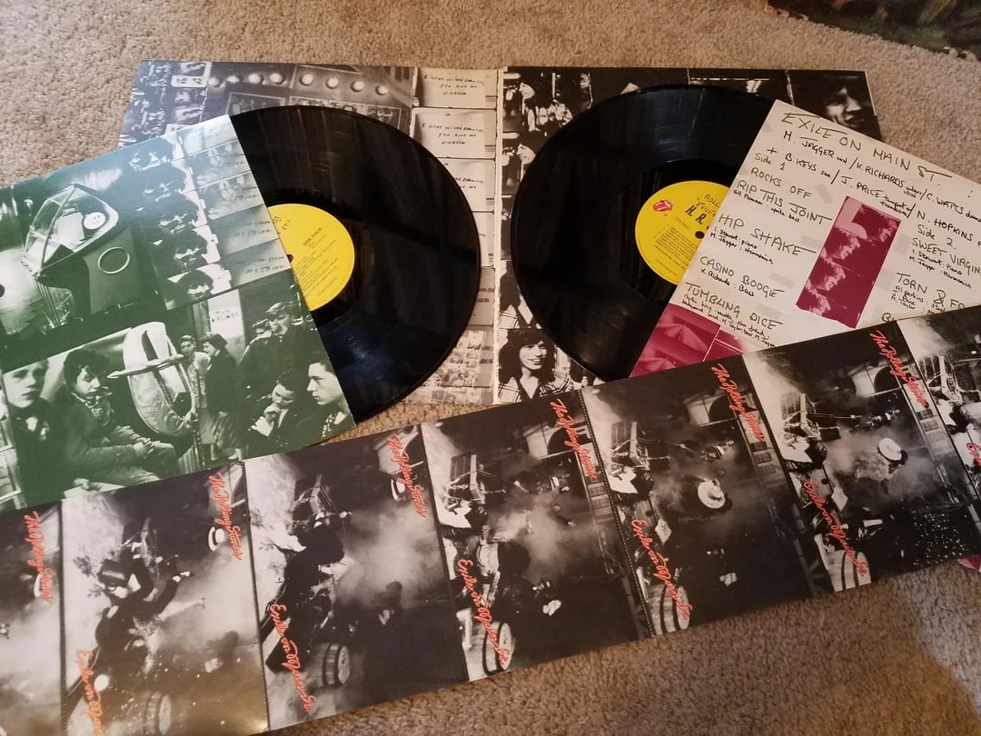 Two vinyl records and their sleeves from The Rolling Stones' album "Exile on Main St." are spread out on a carpet. The covers feature black-and-white photos and handwritten notes; the yellow record labels are visible.