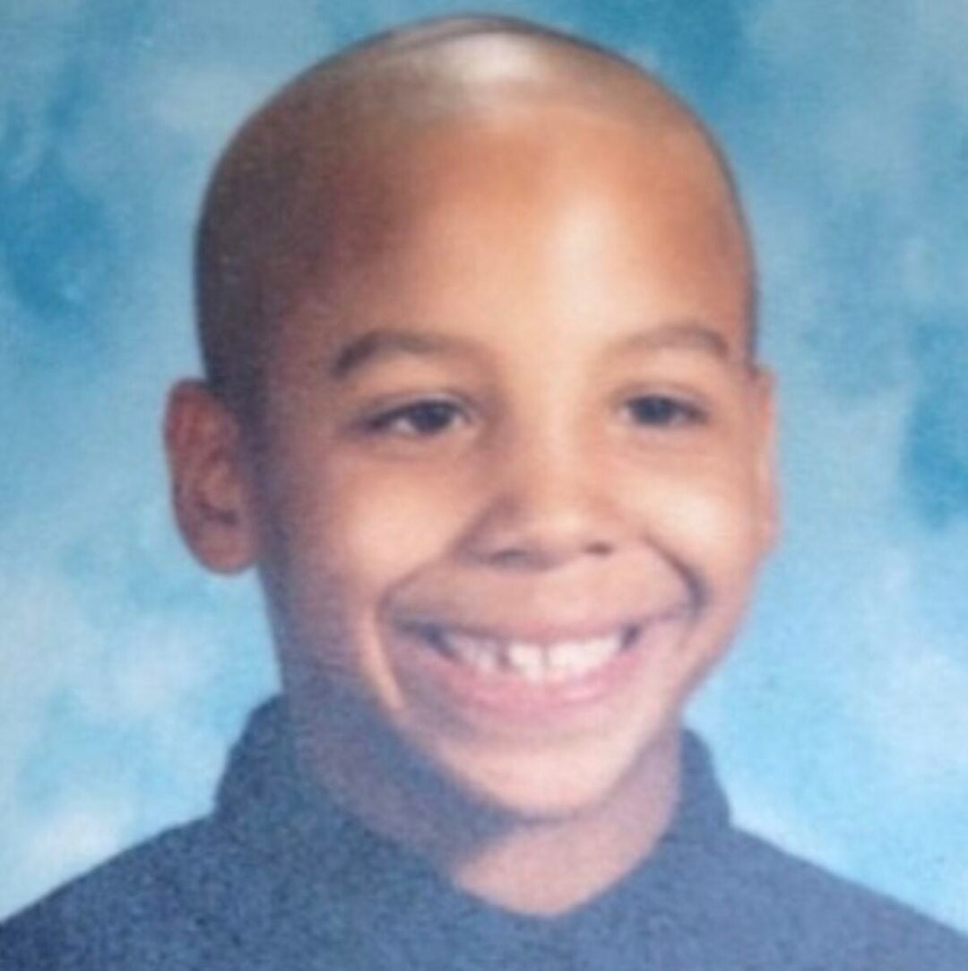 A young child with a shaved head smiles at the camera, wearing a dark shirt against a blue and white mottled backdrop, typical of school portrait photos.