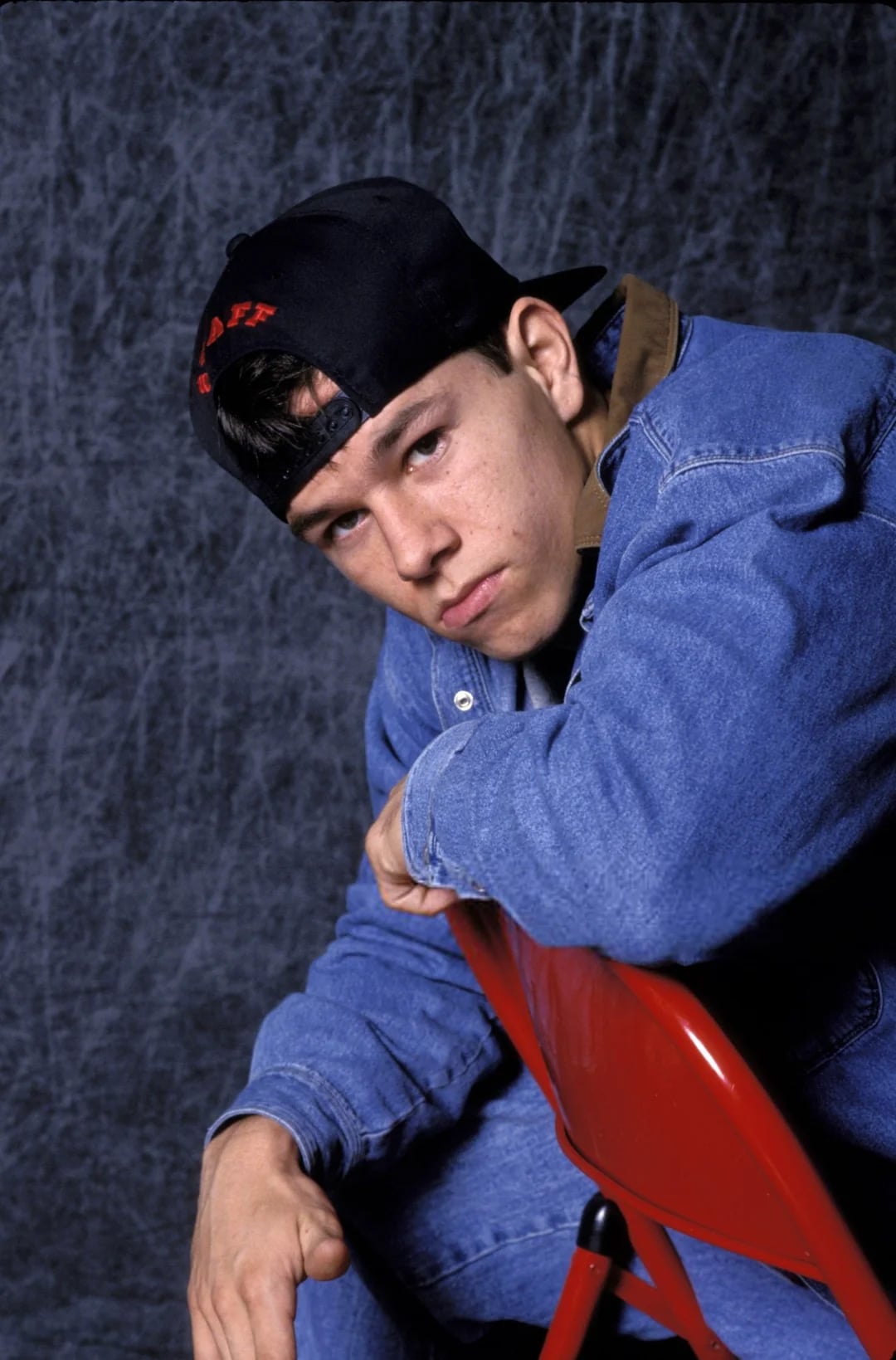 A young man wearing a denim jacket, matching jeans, and a backwards black cap sits on a red chair, leaning forward with a serious expression against a textured dark background.