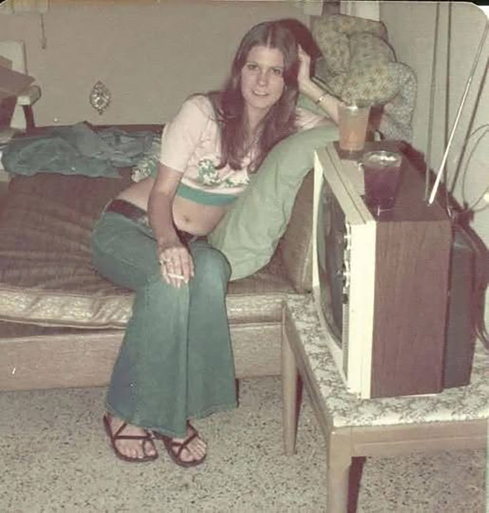 A young woman with long hair, wearing a cropped top and flared jeans, sits on a couch with one arm resting on a pillow. A drink is on the table next to an old-fashioned TV in a retro-styled room.