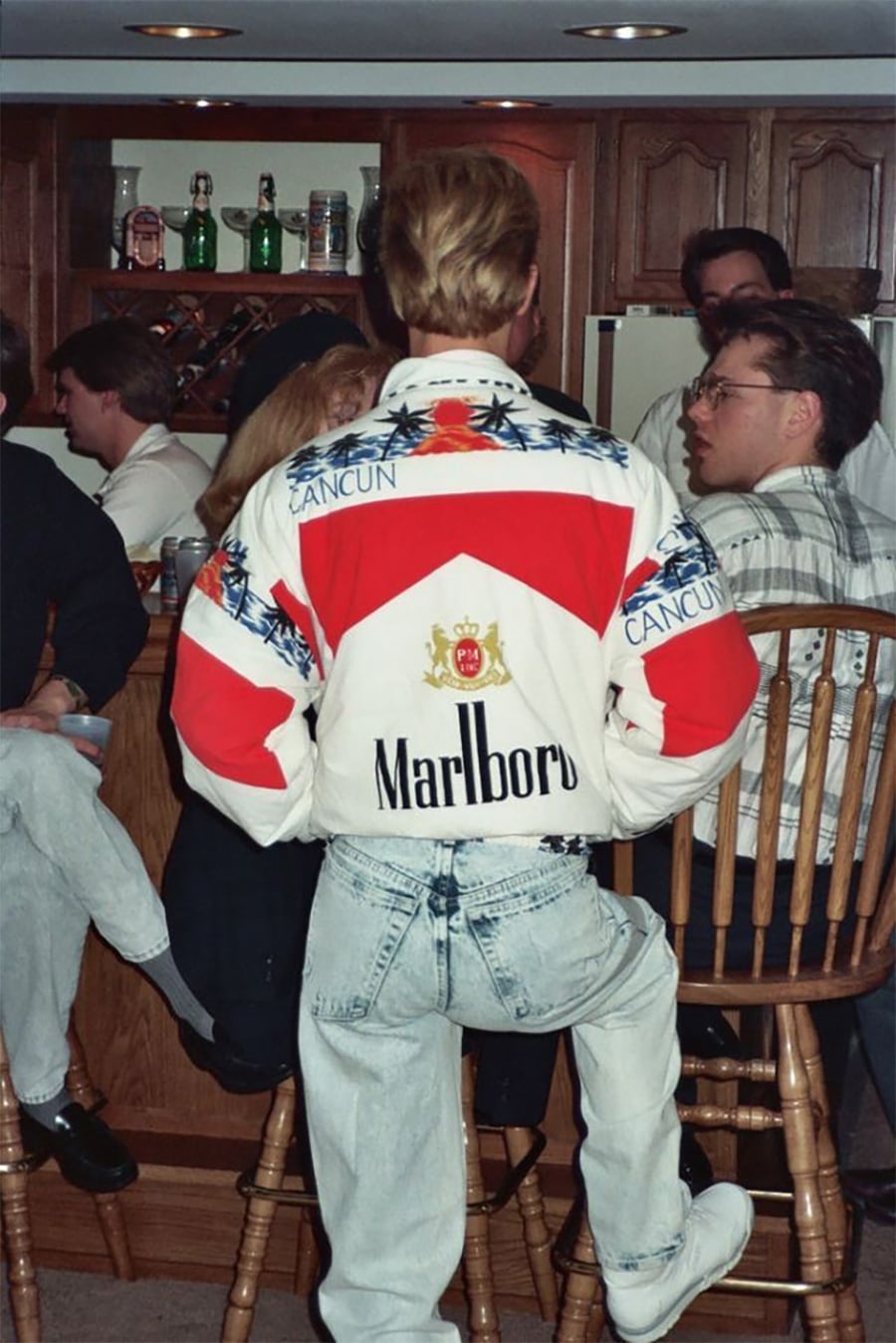 A person with blond hair, seen from behind, wears a colorful "Marlboro Cancun" jacket and acid-wash jeans at a casual indoor gathering with several people sitting around a wooden bar in a kitchen.