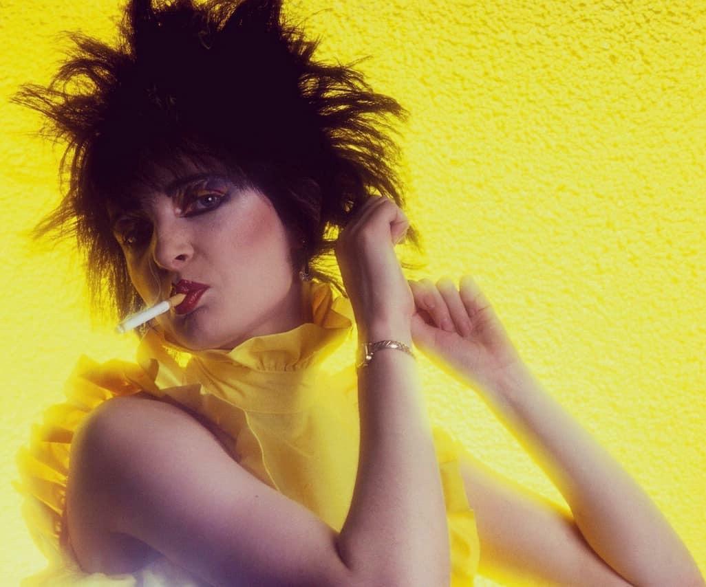 A person with spiky black hair, dramatic makeup, and a ruffled yellow top poses against a bright yellow background, holding a cigarette to their lips.