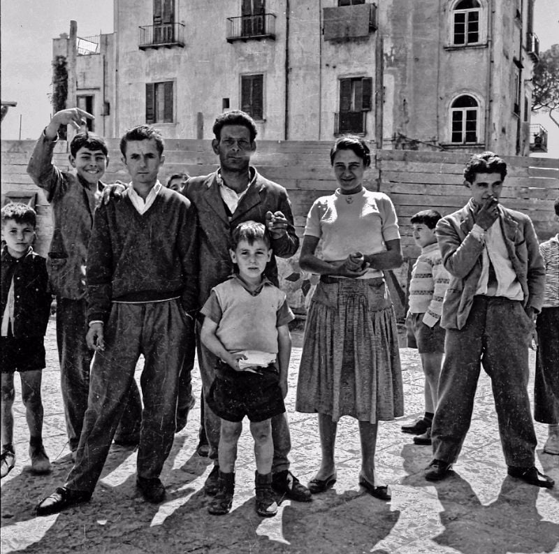 A group of boys and a woman stand outdoors on a stone plaza in front of a large building. Some people look directly at the camera while others pose casually or gesture, creating a lively atmosphere.