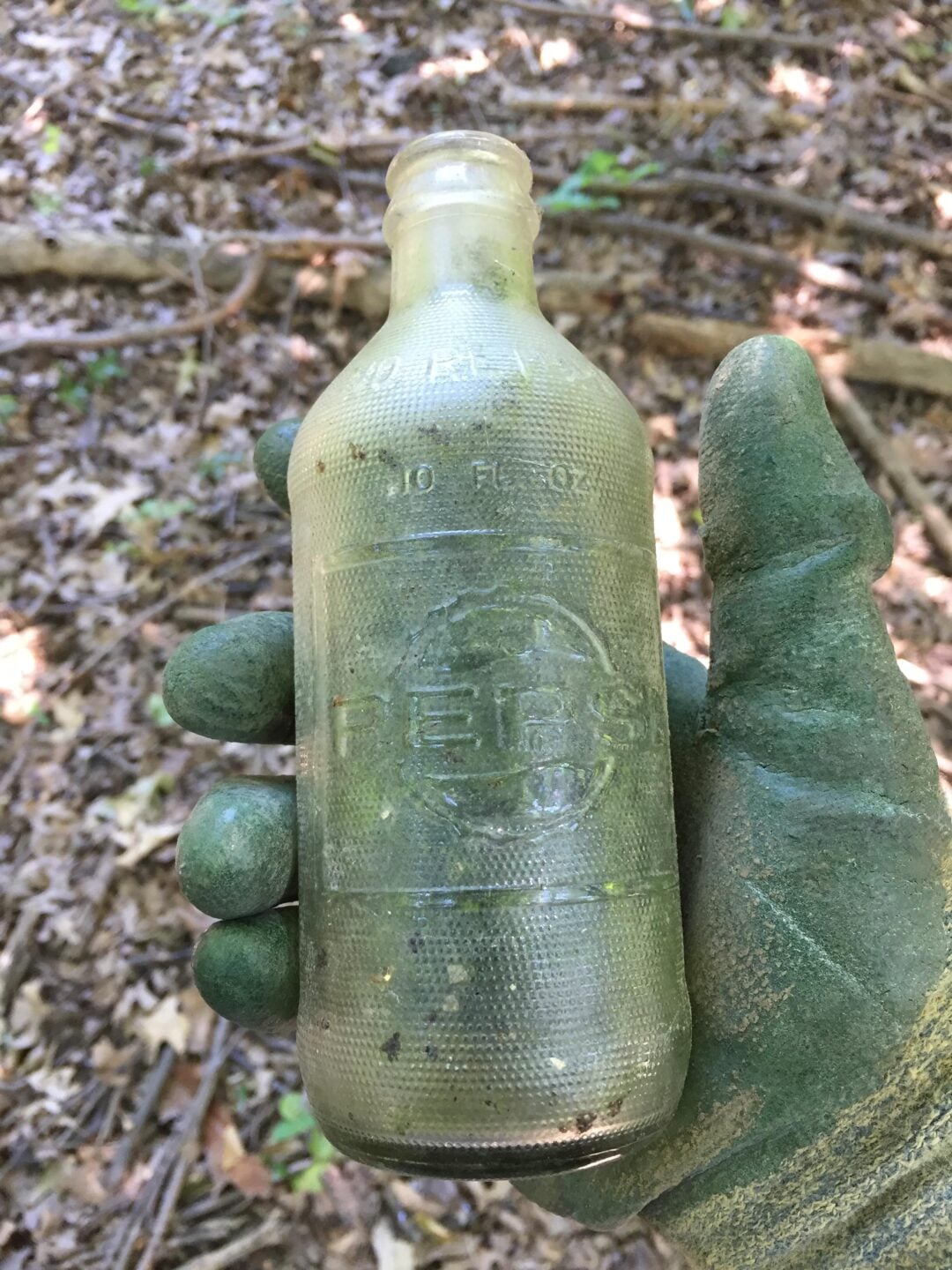 A gloved hand holds an old, dirty glass bottle embossed with the words "10 FL OZ" and a circular logo, with a forest floor covered in dry leaves visible in the background.