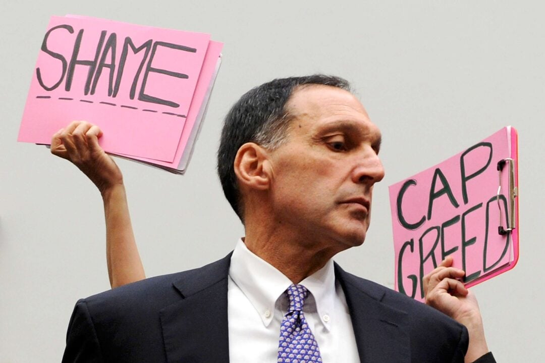 A man in a suit stands in front of two raised pink signs reading "SHAME" and "CAP GREED" during what appears to be a protest or public hearing.