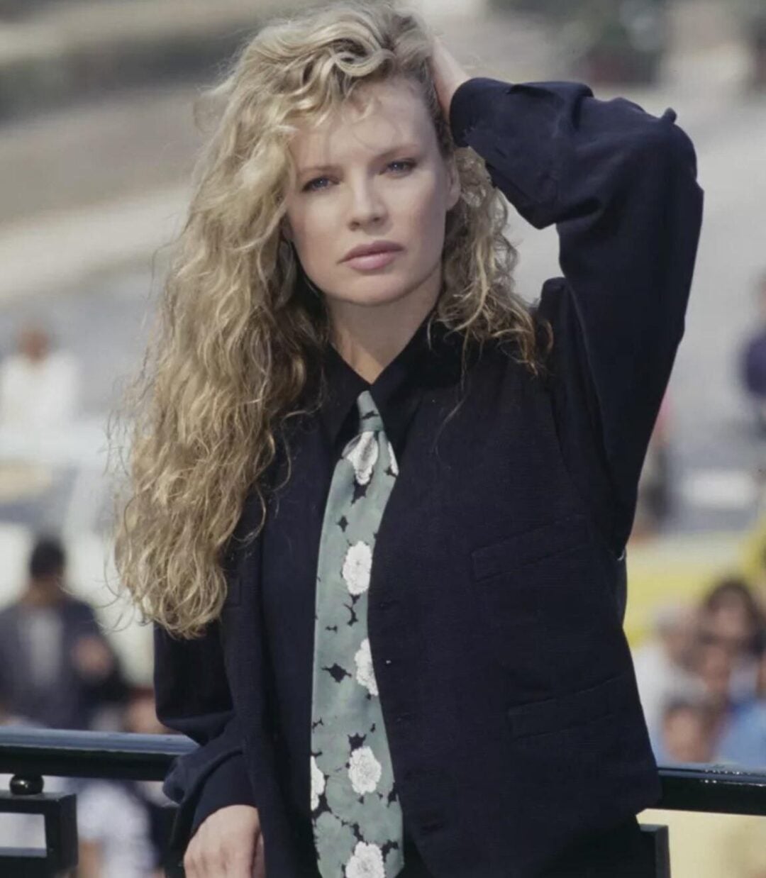 A woman with long, curly blonde hair wears a dark jacket and a floral tie, standing outdoors with one hand resting on her head and a serious expression. The background is blurred with people and a road visible.