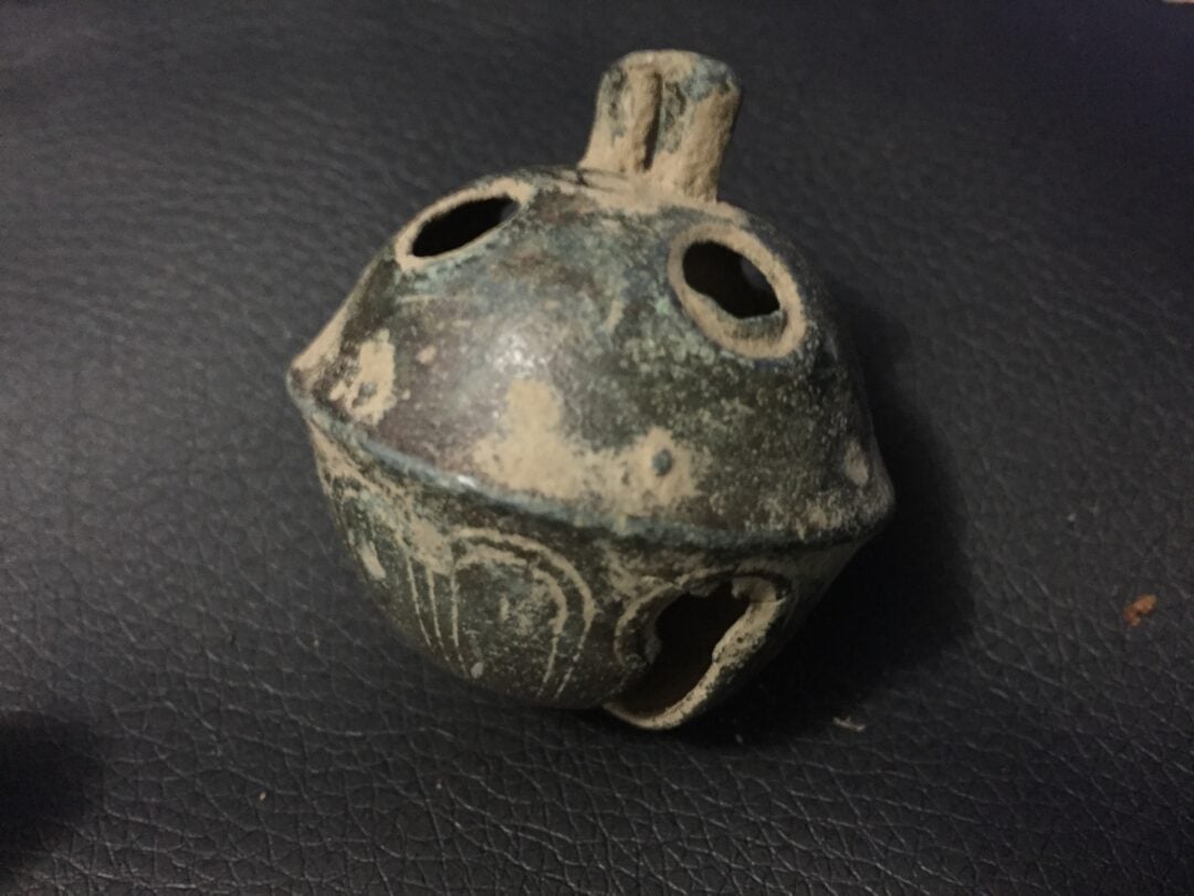 A weathered, round metal object with a seam around the middle, multiple circular holes, and a small stem on top, resting on a textured dark surface.