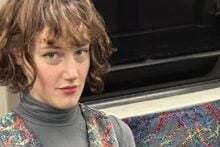 A person with short brown hair wearing a gray turtleneck and a colorful patterned dress sits on a patterned seat in what appears to be a subway or train.
