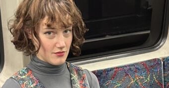 A person with short brown hair wearing a gray turtleneck and a colorful patterned dress sits on a patterned seat in what appears to be a subway or train.